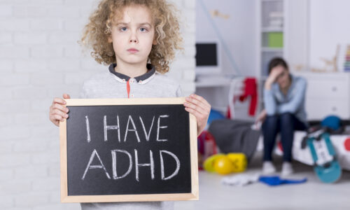 A young boy holding a chalkboard reading "I HAVE ADHD" with a stressed parent in the background