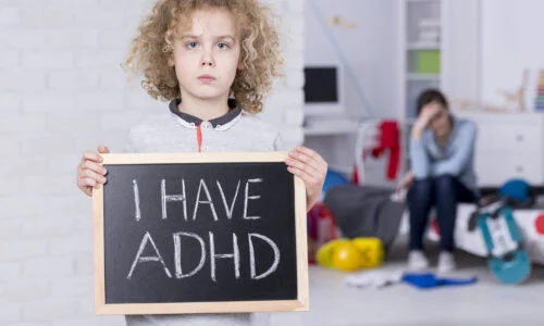 A young boy holding a chalkboard reading "I HAVE ADHD" with a stressed parent in the background